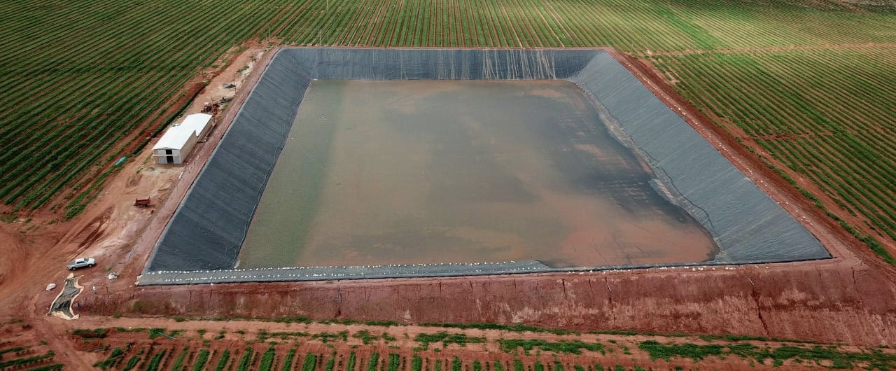 Vista aérea de reservatório agrícola revestido com geomembrana, cercado por lavoura